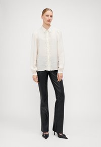 Blouse de couleur crème avec un col, tissu texturé, boutons décoratifs sur le devant, et manches bouffantes longues, associée à un pantalon en cuir noir.