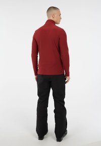 Maglione rosso a maniche lunghe con collo alto realizzato in tessuto morbido, abbinato a pantaloni neri. Presenta un design aderente e un colletto leggermente allungato.