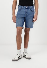 Shorts din denim de culoare bleu deschis, cu un croi clasic, având cinci buzunare și margini răsucite, asortați cu adidași gri și alb.