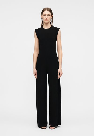Femme debout portant une combinaison noire sans manches avec un pantalon jambes larges, des cheveux longs et raides, des boucles d'oreilles pendantes, et des talons ouverts noirs.
