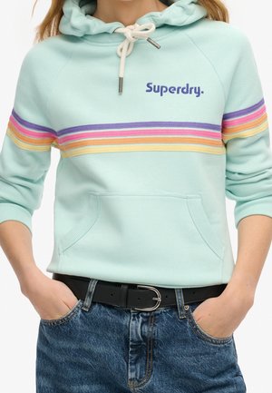 Hellblauer Hoodie mit Regenbogenstreifen über der Brust, "Superdry" Logo links, weiße Kordel, getragen mit blauen Jeans und schwarzem Gürtel, Hände in den Taschen.