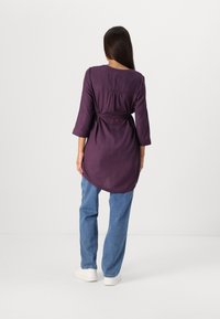 Tunic viola a maniche lunghe con vita regolabile, scollatura rotonda e pieghe sul retro, abbinata a jeans azzurri e sneakers bianche.