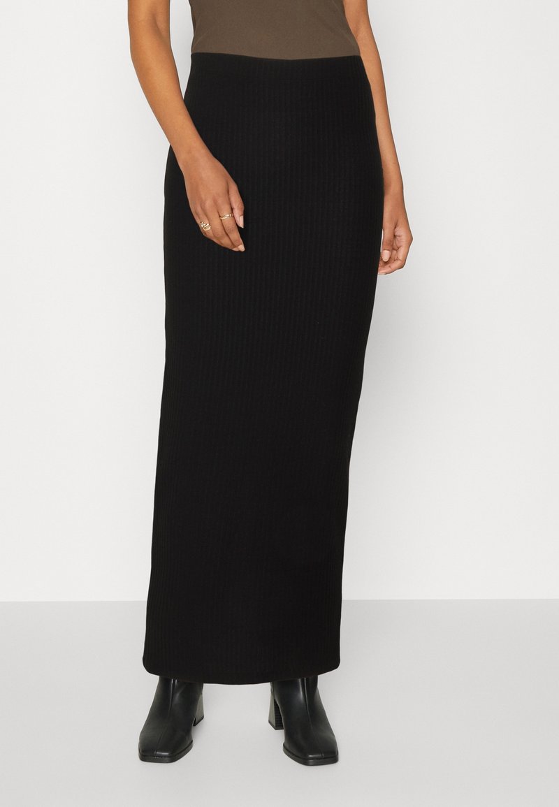 Jupe maxi côtelée en tissu noir, silhouette cintrée, taille haute, texture verticale, associée à des bottines noires.