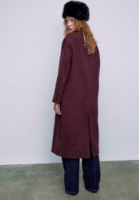 Bordeaux uldfrakke med lige snit, sideslidser og knapdetaljer. Kombineret med mørke brede jeans og en sort pels hat.