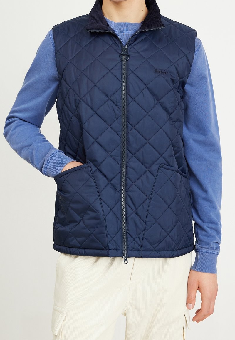 Marineblau gesteppte Weste mit hohem Kragen, Reißverschluss vorne und zwei Seitentaschen. Besitzt eine weiche Textur und kontrastierende Cordura-Akzente am Kragen.