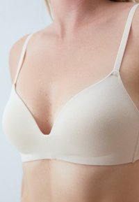 Beige seamless melltartó, mély V-nyakkivágással, állítható pántokkal és puha textúrával. Nincs látható hardver vagy minta.