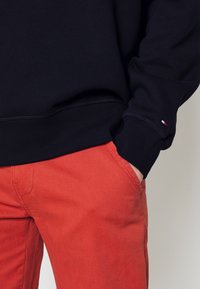 Sudadera negra con cintura acanalada, combinada con pantalones rojo-naranja que cuentan con un bolsillo lateral y costuras visibles. Pequeño detalle de logo en la manga.