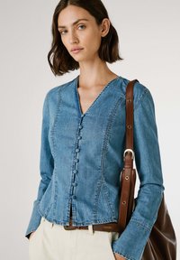 Ljusblå denimjacka med lång ärm, V-ringning och knäppt front. Har sömnadsdetaljer och en skräddarsydd passform. En brun rem från en väska är synlig.