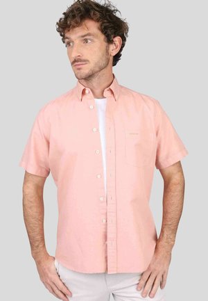 Chemise à manches courtes en tissu rose clair avec un col boutonné, une poche poitrine et un design uni. Ourlet légèrement incurvé et boutons visibles.