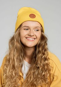 Żółta, prążkowana czapka beanie z okrągłą brązową skórzaną łatą. Nosi ją osoba w jasnej bluzie, prezentując długie, falujące blond włosy.