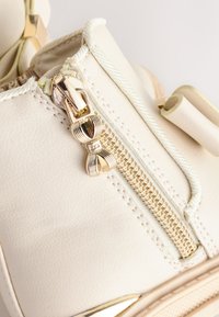 Baskets synthétiques couleur crème avec une fermeture éclair métallique dorée, des coutures détaillées et une surface texturée. Elles présentent un bout arrondi et des accents de lacets.