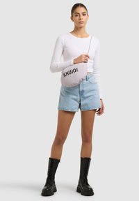 Frau trägt ein weißes Langarmshirt, helle Jeansshorts, schwarze Schnürstiefel und hält eine hellgraue Schultertasche der Marke KHUJO.