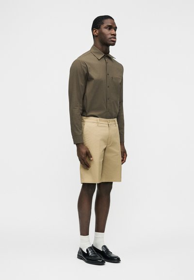 Homme portant une chemise olive à manches longues avec boutons, un short beige jusqu'aux genoux, des chaussettes blanches et des mocassins noirs, debout devant un fond clair.