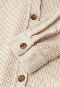 Beige korduroy košile s vertikálním ribbovaným vzorem, s hnědými knoflíky a ohnutým manžetou, zdůrazňující detaily šití.