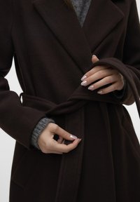 Cappotto di lana marrone con colletto a bavero, cintura in vita e tasche laterali. La mano del modello aggiusta la cintura, mostrando un maglione grigio a maglia sottostante.