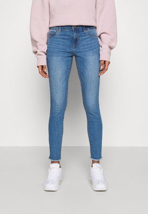 Person iført lyseblå skinny jeans med frynsete kanter, hvide sneakers og en lyserød langærmet top, der står mod en ensfarvet baggrund.