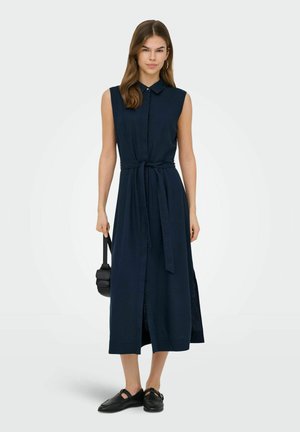 Mujer con vestido midi azul marino sin mangas con cuello y cinturón en la cintura, llevando un pequeño bolso negro y calzando zapatos planos negros.