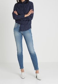 Chemise bleu marine boutonnée avec des pois blancs, associée à un jean slim bleu clair et des mules blanches à bout pointu. Tissu lisse et coupe ajustée.