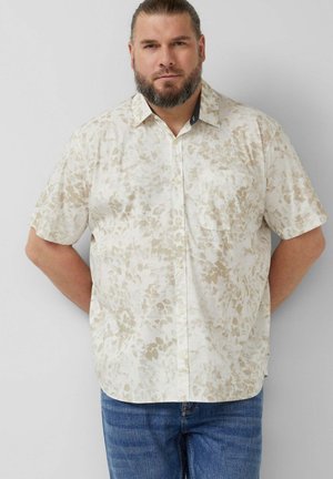 Bärtiger Mann mit zurückgebundenem Haar, der ein beige-weißes, kurzärmeliges Hemd mit Blumenmuster und blaue Jeans trägt und mit den Händen hinter dem Rücken steht.