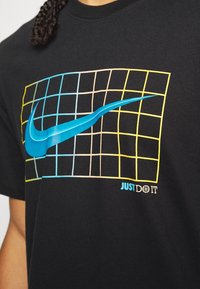 Camiseta de algodón negra con un gran logo de Nike en azul sobre un diseño de cuadrícula en colores amarillos y pasteles, con "JUST DO IT" en texto azul.