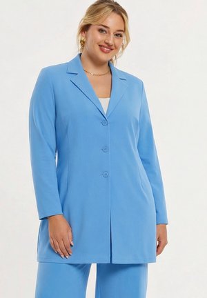 Frau mit blonden Haaren, die goldene Creolen und ein hellblaues Sakko mit passenden Hosen trägt, leicht lächelnd.