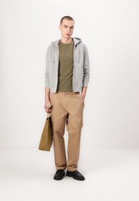 Felpa grigia con zip sopra una maglietta verde oliva, abbinata a pantaloni beige. Scarpe nere, mentre tiene una borsa a rete beige. Design minimalista, vestibilità casual.