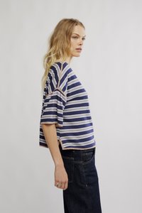 Srajca s kratkimi rokavi in črtami v modri in krem barvi. Mehka tekstura, ovratnik okrogle oblike in vidni šivi. Nošena s temnimi kavbojkami.