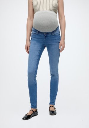 Tehotná osoba oblečená v sivom tehotenskom tielku, modrých úzkych tehotenských džínsoch a čiernych balerínach Mary Jane, stojaca na bielom pozadí.