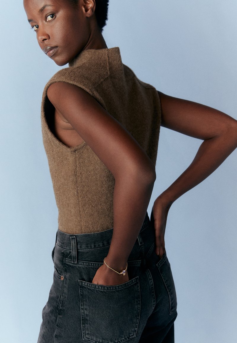 Brun ärmlös stickad topp med hög hals, i kombination med högmidjade svarta jeans. Modellen står med en hand på höften. Mjuk bakgrund.