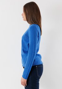 Maglione blu lavorato a maglia con maniche lunghe e scollo rotondo. Tessuto morbido e design aderente, abbinato a jeans denim scuri. Vista laterale.