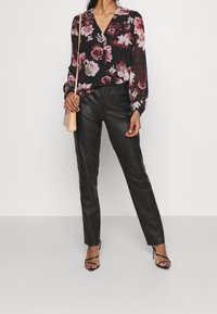 Zwarte bloes met bloemenprint en lange mouwen, gecombineerd met hoge zwarte leren broeken. Eenvoudige sandaaltjes met een bandje en een kleine, gestructureerde roze tas maken de look compleet.