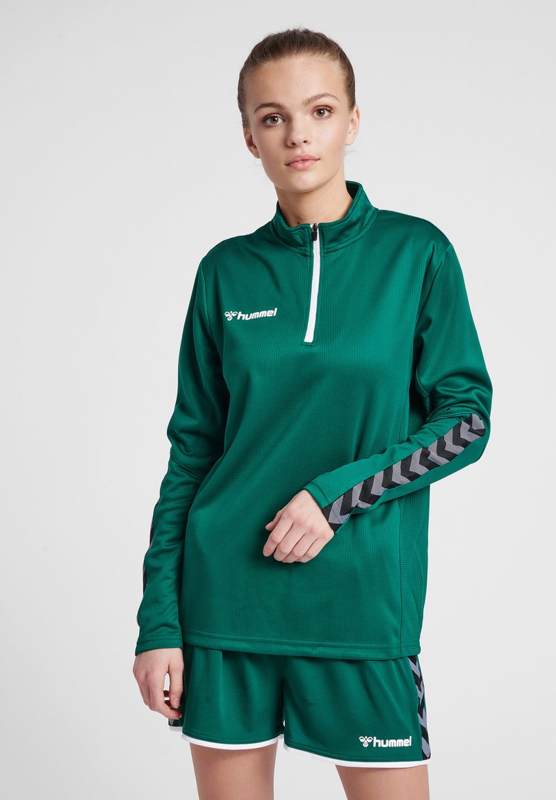 Maglia sportiva verde con zip e tessuto testurizzato, caratterizzata da un logo bianco e dettagli a chevron grigi sulle maniche. Abbinata a pantaloni corti coordinati.
