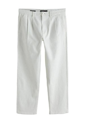 Mann trägt blaues Langarmhemd, weiße Hose, Sonnenbrille, steht drinnen nahe einer beigen Wand mit geflochtenem Korb auf dem Boden.