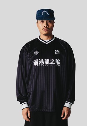 Mand iført en sort Hong Kong Dragons sportsjersey og en blå kasket med hvidt fjer-design står foran en ensfarvet baggrund.