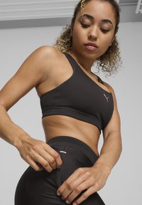 Juodas sportinis liemenukas su asimetriniu dizainu ir logotipu, derinamas su juodomis aukšto liemens leggings'ais, turinčiais mažą kišenę ir tinklinę detalę.