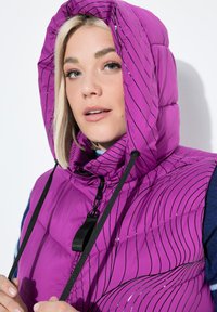 Žena s blond vlasy, oblečená v fialové bundě s kapucí s černými pruhy, drží šňůrky od kapuce na bílém pozadí.