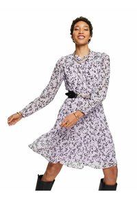 Vestido de flores moradas con mangas largas y dobladillo con volantes. Estilizado con un cinturón negro y usado con botas negras altas.