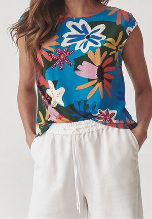 Bezrekawnik w jaskrawym niebieskim kolorze z kolorowymi dużymi kwiatowymi wzorami, noszony z luźnymi białymi spodniami ze sznurkiem.