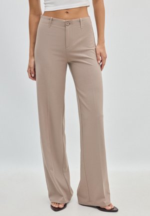 Femme portant un pantalon beige taille haute à jambes larges avec fermeture bouton et zip, assorti à des sandales noires ouvertes aux orteils et un crop top blanc.