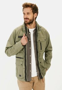 Veste vert olive avec plusieurs poches, fermetures à pression et texture douce. Portée par-dessus une chemise grise, avec un design intérieur contrasté.