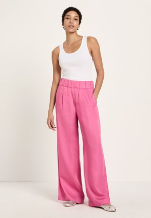 Woman standing wearing a white sleeveless top, high-waisted loose pink pants, and silver ballet flats on a plain background.