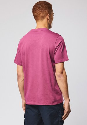 Man with short curly hair wearing a plain pink short-sleeve t-shirt and dark navy cargo pants, standing against a light gray background.