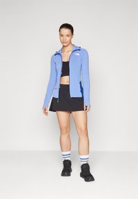 Ljusblå zip-up träningsjacka med långa ärmar, ihopparad med en svart crop top och kjol. Svarta stövlar och randiga strumpor fulländar looken.