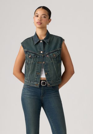 Jeune femme aux cheveux foncés porte une veste en denim sombre sans manches par-dessus un crop top blanc et un jean bleu sur un fond clair uni.