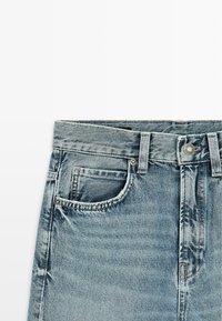 Blauwe spijkerbroek met voorzak, riemlussen en metalen knoopsluiting, zichtbaar op een witte achtergrond.