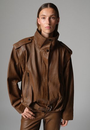 Modèle portant une veste en cuir marron à col montant et ceinturée, assortie d’un pantalon coordonné, sur un fond gris uni.