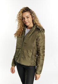 Giacca puffer verde oliva con cappuccio, chiusura con zip, polsini elastici e texture trapuntata. Indossata sopra un maglione leggero con scollatura tonda.