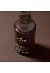 Brun cylindrisk behållare med pumpöppning, märkt "NUXE men." Texten framhäver ögonkrämens användningar: anti-trötthet och anti-mörka ringar egenskaper.