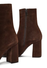 Botines de ante marrón con punta cuadrada, tacón alto tipo bloque y detalle de cremallera lateral. El material presenta un acabado suave y texturizado.