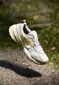 Chaussure de sport beige et grise avec une tige en maille, des accents texturés, des détails réfléchissants et une semelle épaisse, sur fond naturel.